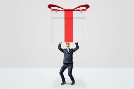 A small businessman holding a giant gift box with both hands above his head.の写真素材