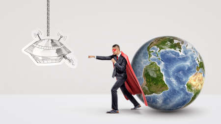 A superhero businessman fighting off a paper drawing of a UFO while behind him stands a small Earth globe.の写真素材