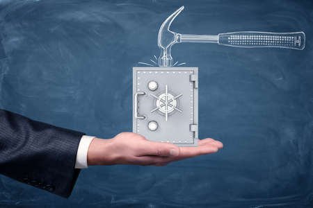 A businessmans hand turned up and a small silver safe standing on it below a drawn hammer on chalkboard background.の写真素材