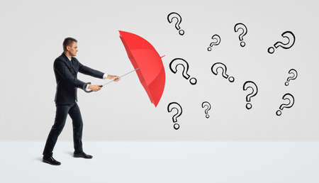 A businessman covering himself with an open red umbrella from many black drawn question marks.の写真素材