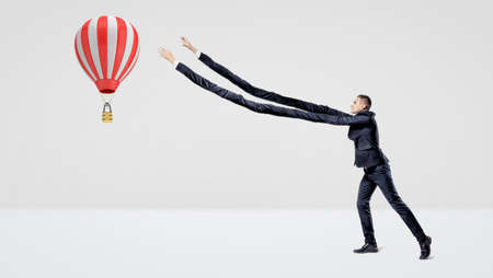 A businessman in side view trying to catch a large flying hot air balloon with his extra-long arms.の写真素材