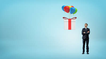 A small businessman looking up at a large gift box flying away on colorful balloons.の写真素材
