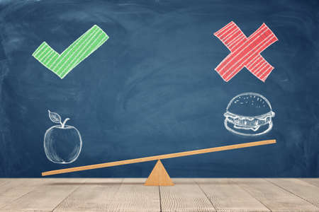 A wooden seesaw on blue chalkboard background where a painted apple overweighs a sandwich.の写真素材