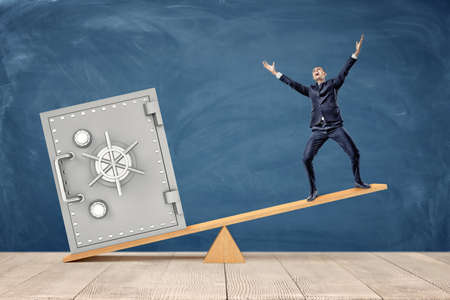A tiny businessman standing on wooden seesaw while being overweighed by a large silver safe box.の写真素材