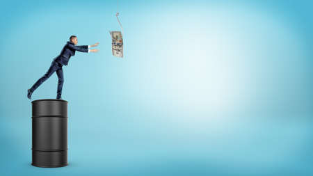 A small businessman standing on a large oil barrel and trying to catch a dollar bill from a silver hook.の写真素材
