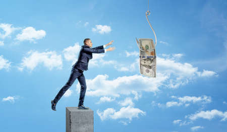 A small businessman standing on a concrete column and catching a dollar bill caught on a metal hook.の写真素材