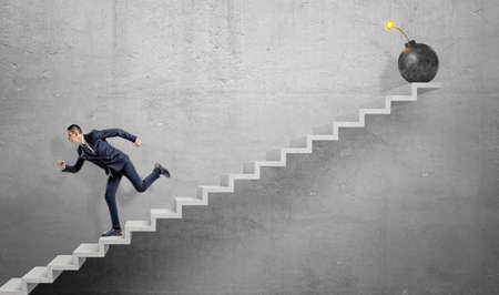 A scared businessman running down grey concrete stairs away from a large iron bomb with a lit fuse.の写真素材