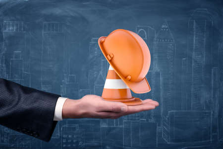 A striped traffic cone and an orange hard hat standing on businessmans palm.の写真素材