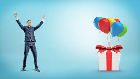 A happy businessman with hands raised up standing behind a gift box tied to many balloons.の写真素材