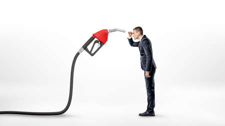 A businessman on white background staring closely at a large red gas nozzle attached to a black...