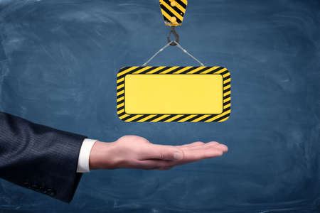 A blank yellow striped construction sign hanging from a crane hook above businessmens palm on chalkboard background.の写真素材