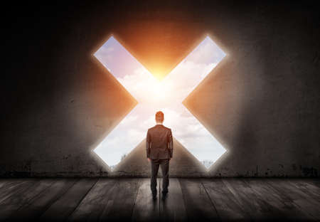 A businessman stands with his turned back and looks into a cross-shaped hole in a concrete wall.の写真素材