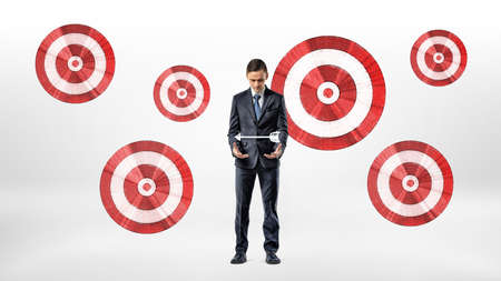 A businessman stands beside a wall with many archery targets and looks at an arrow hovering over his...