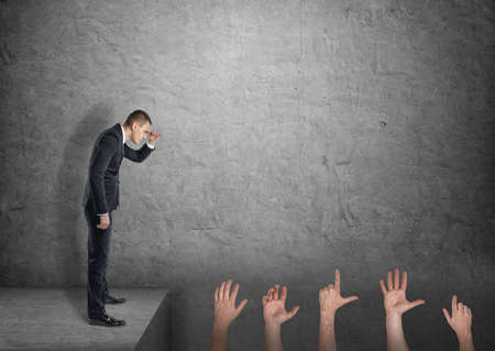 A businessman on grey concrete background looks down into an abyss full of pointing, grabbing and open human palms.の写真素材