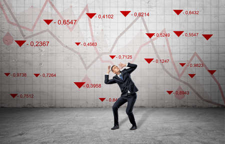 A scared businessman cowers near a concrete wall with red stock market indexes and falling statistic lines.の写真素材