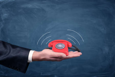 A businessman's palm hold a small red retro dial phone with lines showing soundwave emissions....