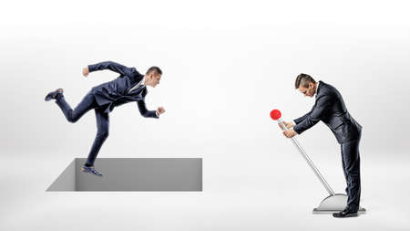 A businessman runs on white ground while a square hole controlled by a man on a lever opens right under his feet.の写真素材