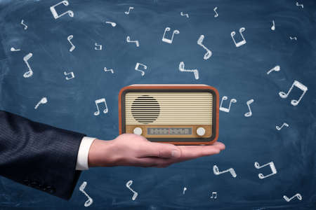 A small retro radio set with brown housing and yellow front stands on a businessmans palm on blackboard background. Broadcasting business. Earn money from podcasting. Radio host.の写真素材