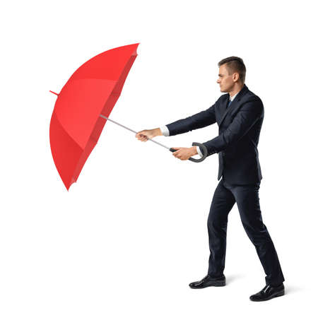 A young businessman stands on white background and hides from harsh environment behind an open red umbrella.の写真素材