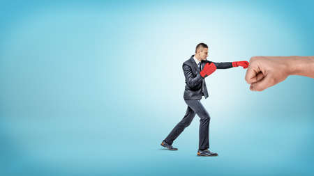 A small businessman in red boxing gloves punches a giant male fist on blue background.の写真素材