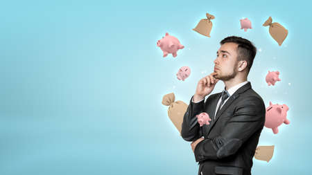 A young bearded businessman deep in thought with small piggy banks and money sacks flying around his head.の写真素材