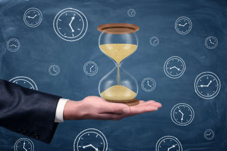 A businessmans hand turned palm up and holds a large functioning hourglass on blue chalkboard...
