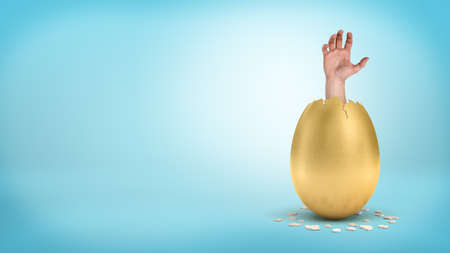 An open male hand sticks out from inside a cracked golden egg on blue background.の写真素材