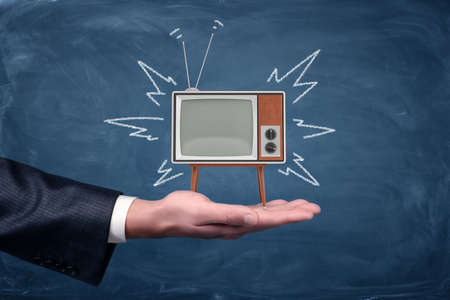 A businessmans palm holds a small retro TV with legs and antenna on blackboard background.