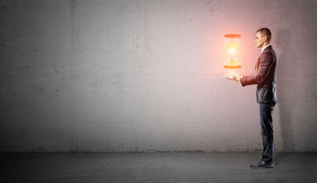 A businessman stands in side view on concrete background with his harms under a hovering glowing hourglass.の写真素材