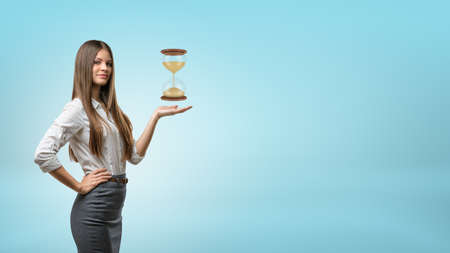 A blond businesswoman stands and holds one hand palm up with a large hourglass hovering above it.の写真素材