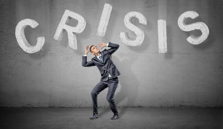 A businessman cowers under large concrete letters making up a word Crisis above him.の写真素材