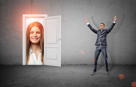 A happy businessman in victory motion stands in a room with a white door opening to a giant happy female face.の写真素材