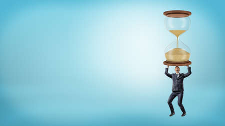A businessman holds a large heavy hourglass in his hands on blue background.の写真素材