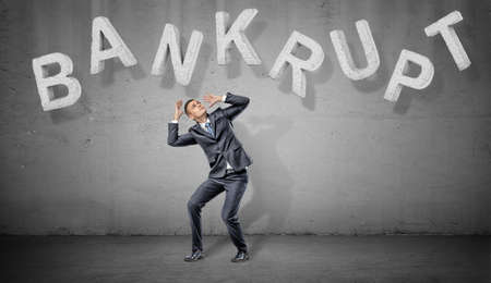 A scared businessman hides under large concrete letters making a word Bankrupt above him on a grey background.の写真素材