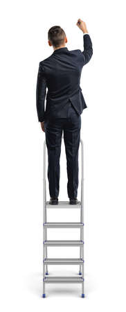 A businessman stands on a metal stepladder and draws with one hand in a back view on a white background.の写真素材