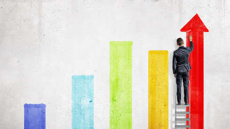 A businessman stands on a stepladder and draws with a paint roller several colorful chart columns.の写真素材