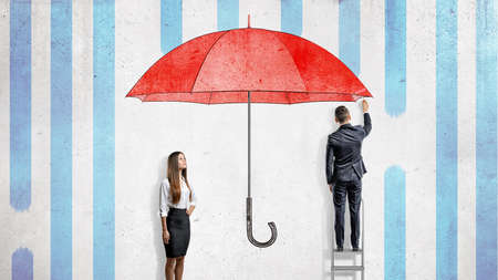 A businesswoman stands near a wall where a businessman draws a giant red umbrella covering them from the rain.の写真素材