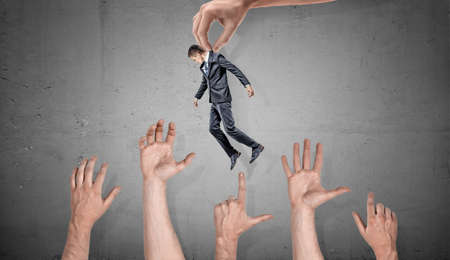 A giant hand holds a small businessman over many giant grabbing hands on concrete background.の写真素材