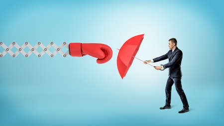 A small businessman holding a red umbrella to hide from a large boxing glove on a spring arm.の写真素材