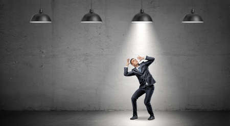 A scared businessman stands cowering under the rays of industrial pendant lights right above him.の写真素材