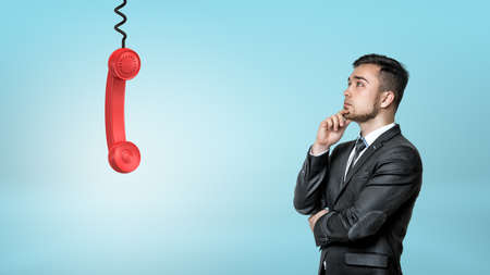 A thinking businessman looks up on a red retro phone receiver hanging from a black cord.の写真素材