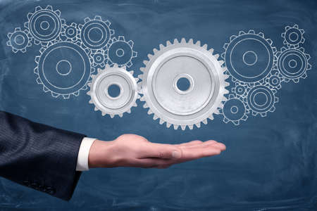 A businessmans hand holds two connected gears on a blackboard background with many other gears drawn on it.の写真素材