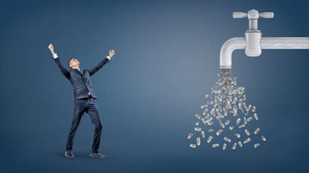 A small victorious businessman stands with raised arms near a giant water faucet leaking a lot of dollar bills.の写真素材