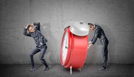 A businessman covers his head form a giant red retro alarm clock being winded up by another businessman.の写真素材