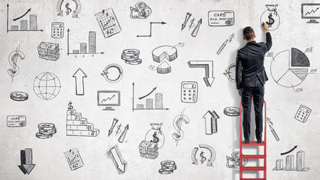 A businessman stands on a red ladder and draws financial illustrations on a while wall.の写真素材
