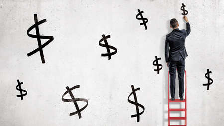 A businessman stands on a red stepladder and draws black dollar signs on a white wall.の写真素材
