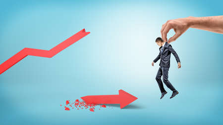 A small businessman is lifted up by a giant human hand on a blue background over a red broken statistic arrow.の写真素材