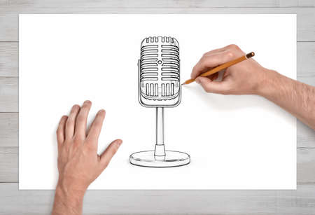 A pair of male hands in close view draws the retro microphone on a short stand with a pencil on white paper.の写真素材
