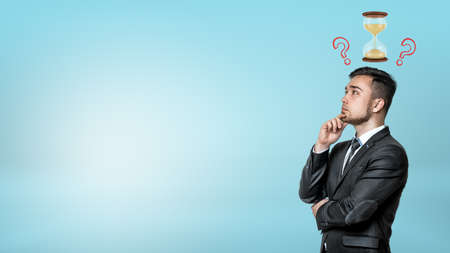 A portrait of a thinking businessman with a small hourglass and question marks above his head.の写真素材