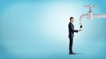 A tiny businessman stands near a giant metal faucet and holds out his hands to catch a giant oil drop.の写真素材